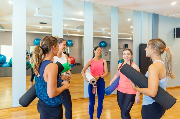 A photo of five women in active wear holding yoga mats talking amongst themselves