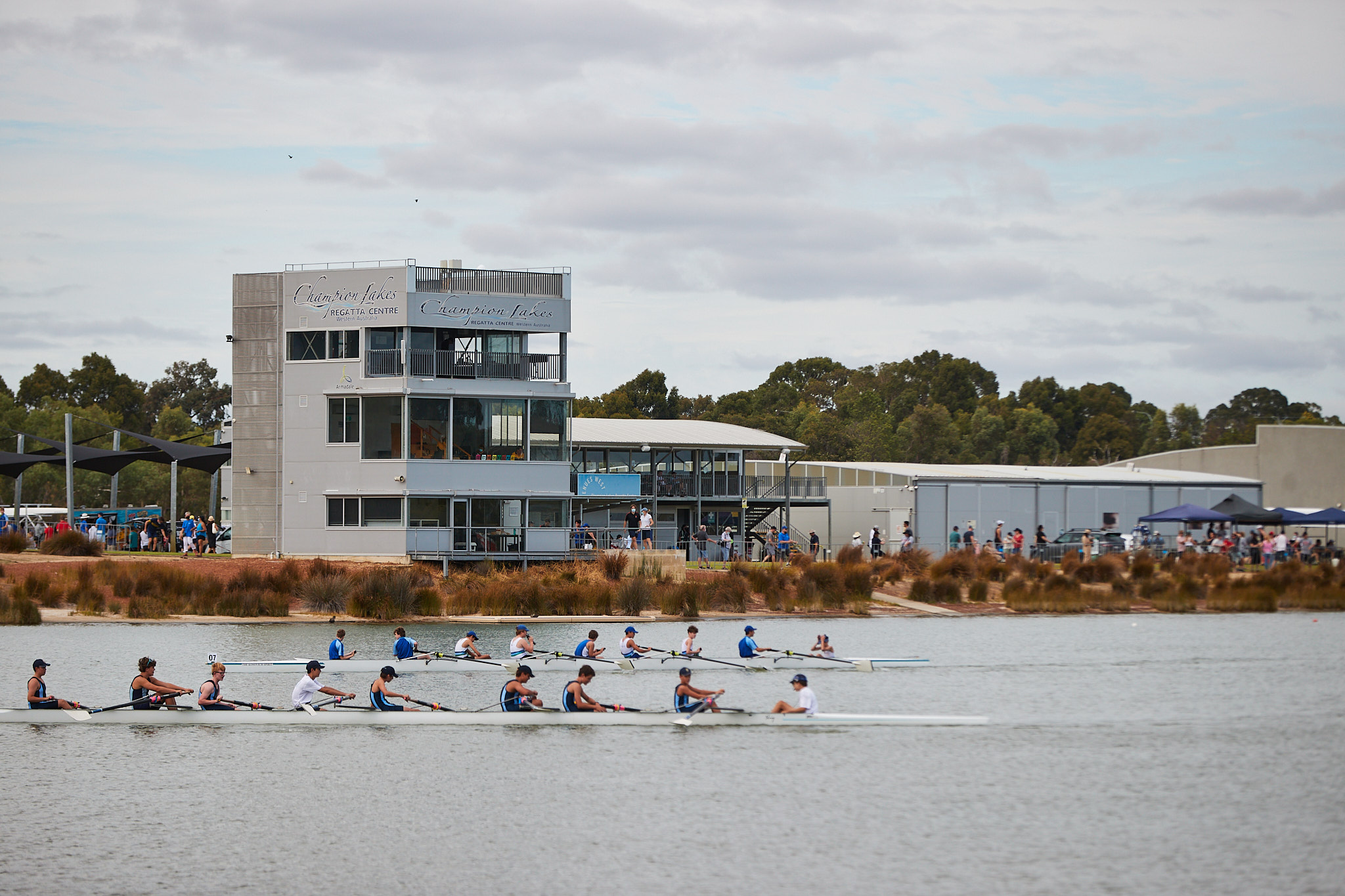 A side oh photo of two rowing teams competing with the main building in the background at the Champion Lakes Regatta Centre in Champion Lakes, Perth