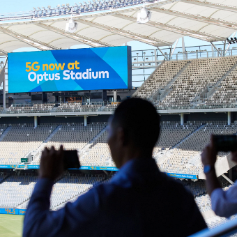 Photo from behind two people with focus on the Optus Stadium big screen in the background
