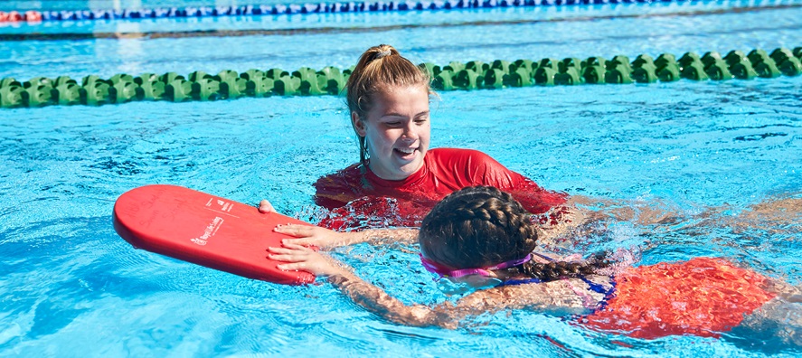 A photo of a swim instructor assisting a child in swimming using a kick board