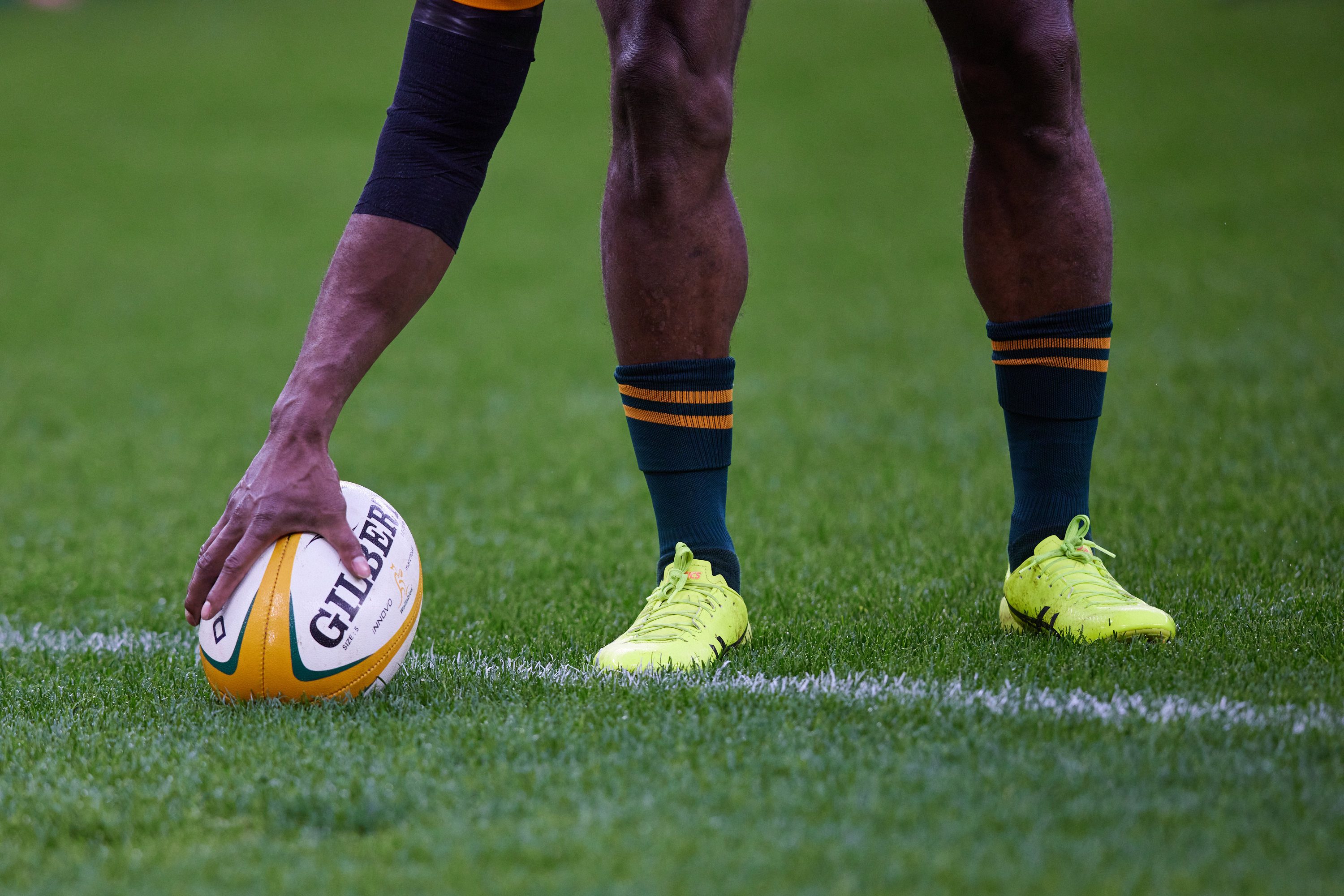 A photo of the lower half of a person bending down to grab a rugby ball on the ground